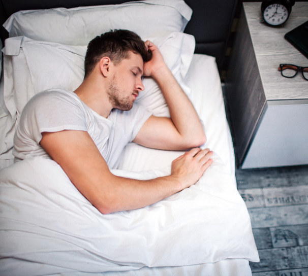 man sleeping on his side with glasses, book and clock on his night stand