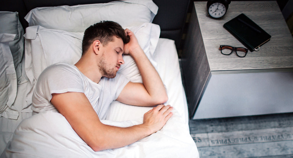 man sleeping on his side with glasses, book and clock on his night stand